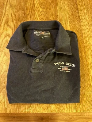 Mörkblå piké från Polo Club - Klassisk mörkblå pikétröja från Polo Club Saint Vincent med vit logga och text på bröstet. Tröjan har krage, knappar vid halsen och är tillverkad i bomull. Perfekt för en avslappnad men stilren look.