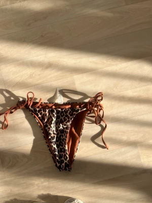 Leopardmönstrad bikiniunderdel med knyt - Snygg bikiniunderdel  med leopardmönster och bruna sidor. Modellen har knyt i sidorna för justerbar passform och är perfekt för sommardagar på stranden.