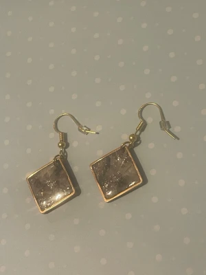 Guldiga örhängen med marmorerad ruta - Unika örhängen med fyrkantig, marmorerad ruta i grått och vitt. Guldfärgade detaljer och krok. Perfekta för dig som vill ha något annorlunda och trendigt i smyckeskrinet. Materialet ser ut att vara metall och resin.