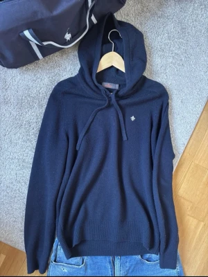 Merinoull hoodie Morris - Snygg mörkblå merinoull hoodie från Morris med huva och dragsko. Diskret logga broderad på bröstet. Tröjan har ribbade muddar och är tillverkad i mjukt stickat material som känns riktigt skönt mot huden. Perfekt för dig som gillar klassisk och stilren design.