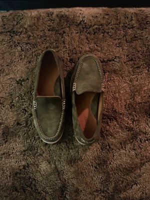 Mörkgröna mocka loafers från Polo Ralph Lauren - Snygga loafers i mörkgrön mocka från Polo Ralph Lauren. Skorna har klassisk design med vita kontrastsömmar och en diskret logga på sulan. Yttersulan är vit med röd Polo-detalj. Perfekta för en stilren look. Passar 41 bra