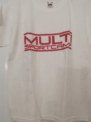 T-shirt - Vit t-shirt från Fruit of the Loom med röd text 'MULTI SPORTCAMP' tryckt framtill och längs ryggen. Klassisk passform med rund hals och korta ärmar. Tillverkad i mjuk bomull, perfekt för sportiga tillfällen eller chill vardagsstil.