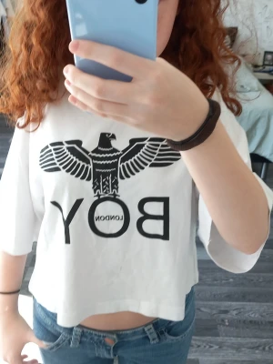 Vit croppad t-shirt från BOY London - Säljer en vit croppad t-shirt från BOY London med ikoniskt svart örntryck och logga på bröstet. T-shirten har korta ärmar och rund halsringning. Perfekt för en edgy och trendig stil.