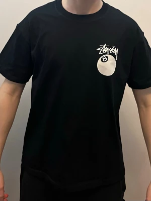 Svart Stüssy t-shirt med 8-ball print - Svart t-shirt från Stüssy med stort vitt 8-ball tryck och logga på ryggen samt mindre print på bröstet. Klassisk passform och rund hals. Tillverkad i mjuk bomull, perfekt för en streetwear-look.