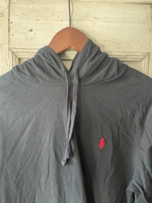 Svart hoodie från Ralph Lauren - Svart tunn hoodie från Ralph Lauren med klassisk röd broderad logga på bröstet. Hoodien har dragsko i huvan och är tillverkad i ett mjukt, tunt material. Perfekt för en avslappnad och stilren look. Ai gjorde beskrivningen 
