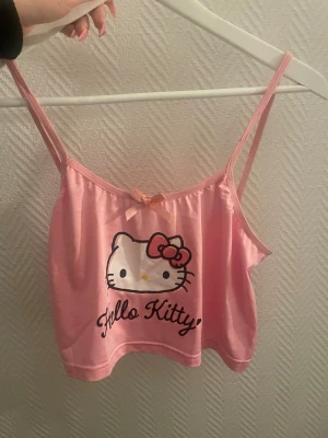 Rosa Hello Kitty linnetopp - Söt rosa linnetopp med Hello Kitty-tryck framtill och liten rosett vid urringningen. Tunn och mjuk med smala axelband, perfekt för en lekfull och trendig look. Croppad modell som passar till både jeans och kjol.