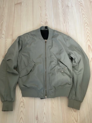 Bomberjacka  - Stilren grågrön bomberjacka med klassisk passform, ribbade muddar vid ärmar och midja samt två snedställda fickor framtill. Jackan har en metallfärgad dragkedja och minimalistisk design utan synligt märke. Perfekt för en clean och avslappnad look.