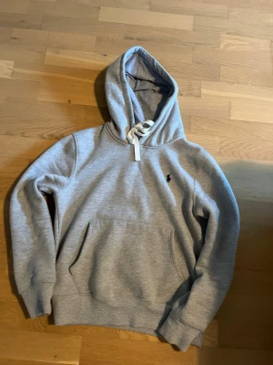 Grå hoodie från Polo Ralph Lauren - Ganska liten i storlek, har krympt i tvätten tror jag. Passar dig runt 170-175. Snygg grå hoodie från Polo Ralph Lauren med klassisk huva och vit dragsko. Mjuk bomullsblandning och känguruficka framtill. Liten broderad logga på bröstet. Perfekt för en chill och stilren look.