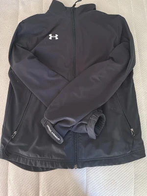 Svart vindjacka från Under Armour - Snygg svart vindjacka från Under Armour med dragkedja framtill och två sidofickor med dragkedja. Jackan har hög krage och diskret logga på bröstet. Tillverkad i ett lätt och vindtätt material som passar perfekt för aktiva dagar utomhus.