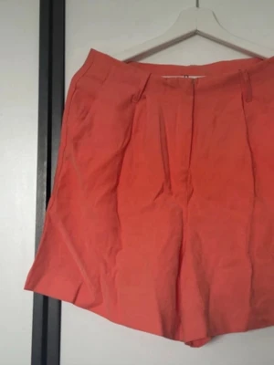 Koralorange shorts storlek 40 - Snygg koralorange skort med hög midja och dragkedja fram. Skorten har en loose passform och är tillverkad i ett lätt tyg. Perfekt för dig som vill ha en chill men ändå trendig look.
