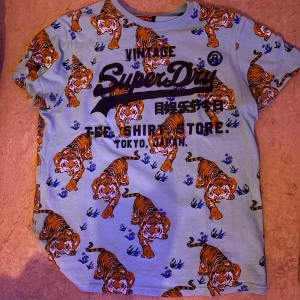 Superdry t-shirt med tigertryck - Cool ljusblå t-shirt från Superdry med stora tigertryck över hela plagget och svart logga framtill. T-shirten har korta ärmar och är gjord i mjuk bomull, perfekt för dig som vill sticka ut med ett unikt mönster.