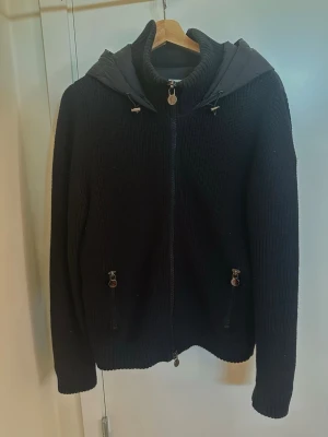 Svart stickad cardigan med huva - Snygg svart stickad cardiganjacka med dragkedja och vadderad huva. Detta är även ett unikt plagg från moncler som är svår att hitta speciellt i bra skick och den passar bra till allt även att man sticker ut och den ser extremt bra ut