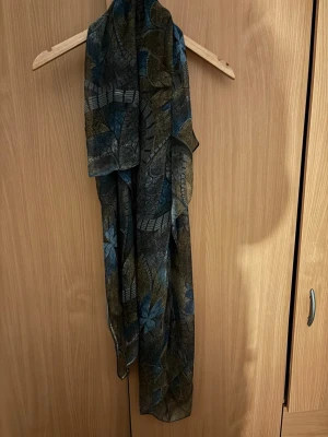 Mönstrad sjal i blått och olivgrönt - Säljer en tunn sjal med bladmönster i blått, olivgrönt och grått. Sjalen har rektangulär form och är lätt transparent, perfekt att slänga över axlarna för en cool look. Materialet känns mjukt och följsamt, och mönstret ger en unik touch till din outfit.