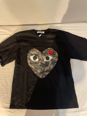Svart Comme des Garçons PLAY t-shirt - Svart t-shirt från Comme des Garçons PLAY med ett stort hjärta i camouflage på bröstet och en liten röd hjärtdetalj. Klassisk rund hals och korta ärmar. Perfekt statement-plagg för dig som gillar streetwear och coola prints.