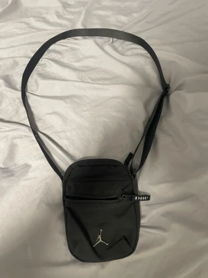 Svart axelväska från Jordan - Snygg svart axelväska från Jordan med det klassiska Jumpman-loggan framtill. Väskan har justerbar axelrem och ett praktiskt ytterfack med dragkedja. Tillverkad i slitstarkt tygmaterial, perfekt för att bära det viktigaste.