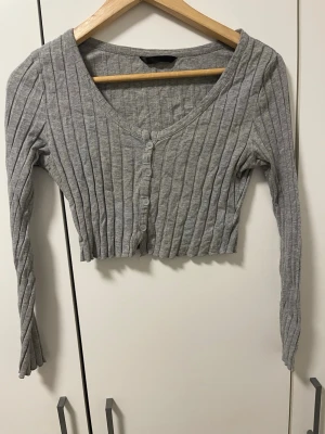 Grå ribbad cropped kofta - Säljer en grå ribbad kofta med knappar framtill och lång ärm. Modellen är cropped och har en djup v-ringning. Perfekt att styla över ett linne eller t-shirt. Materialet känns mjukt och stretchigt.