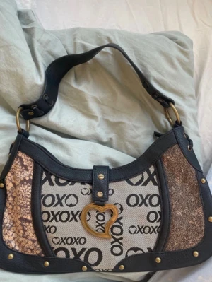 Super snygg vintage 2000-tals väska - Trendig vintage  axelväska från 2000 talet med svart läder, beige tyg med XOXO-logga, ormmönstrade och glittriga paneler samt guldfärgade detaljer. Väskan har en unik hjärtformad metallspänne framtill och bred axelrem med nitar. Passar perfekt för att piffa upp en outfit lite extra