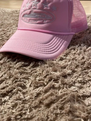 Rosa corteiz keps med fluffig framdel - Säljer en ljusrosa keps med mesh på sidorna och bak, samt broderade vita stjärnor och ett ovalt motiv framtill. Kepsen har böjd skärm och justerbar snapback. Perfekt för dig som vill sticka ut med en trendig accessoar.!UNISEX! 