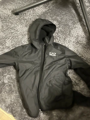 Svart vindjacka från EA7 Emporio Armani - Snygg svart vindjacka från EA7 Emporio Armani med huva och dragkedja framtill. 