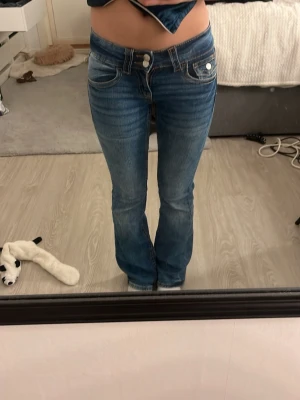 Low waist blå bootcut jeans med dubbla knappar - Säljer ett par blå bootcut jeans med dubbla knappar i midjan och klassiska fem fickor. Jeansen har en snygg tvätt med lätt slitning och kontrastsömmar. Perfekta för dig som gillar en retroinspirerad look och vill ha ett par jeans som sitter snyggt över höfterna. Använt några gånger förra året men har tröttnat på dom. Jag ramlade i gräset sommar 2025 så på högra knät så är det lite mörkare fläck än på det andra, men det tänker man inte på. Passar mig bra som har xxs-xs 