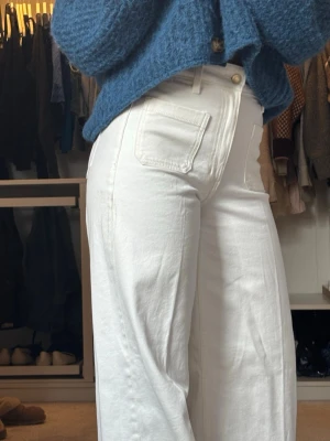 Vita vida jeans - Säljer ett par vita jeans med hög midja och raka, vida ben. Jeansen har stora fickor både fram och bak samt bälteshällor. Perfekta för en clean och trendig look. Materialet känns mjukt och följsamt, troligtvis bomull eller bomullsblandning.