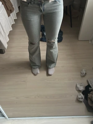 Ljusblå bootcut jeans med slitningar - Säljer ett par ljusblå bootcut jeans med slitningar vid knäna och en röd broderad detalj på bakfickan. Jeansen har hög midja och klassisk femficksdesign. Perfekta för en avslappnad och trendig look.