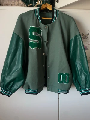 Grön bomberjacka med S och 00 - Snygg grågrön bomberjacka med gröna ärmar i skinnimitation och stora applikationer: ett S på bröstet och 00 nedtill. Jackan har randiga muddar i grönt, vitt och svart samt tryckknappar framtill. Perfekt för en sportig och trendig look.