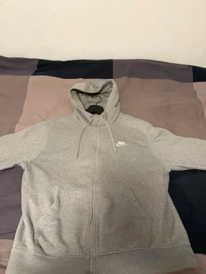 Nike hoodie - Snygg grå zip hoodie från Nike med klassisk logga broderad på bröstet. Hoodien har dragkedja framtill, två fickor och justerbar huva med snören. Tillverkad i mjukt sweatshirtmaterial som är perfekt för chill eller träning.