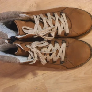 Bruna snörade kängor med ullfoder - Snygga bruna kängor med beige snörning och varmt ullfoder. Kängorna har en klassisk rund tå och robust sula, perfekta för kyligare dagar. Utsidan är i brunt, veganskt läder och insidan har mjukt, ljust foder. Storlek 41.