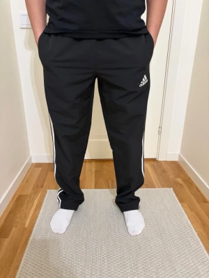 Svarta Adidas mjukisbyxor med vita ränder - Säljer ett par svarta mjukisbyxor från Adidas med klassiska vita ränder längs sidorna och Adidas-logga på vänster lår. Byxorna har rak passform och resår i midjan. Perfekta för chill eller träning.