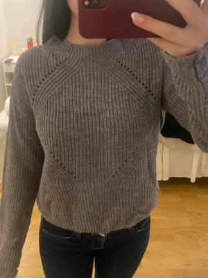Grå stickad tröja med mönster - Mysig grå stickad tröja med diskreta hålmönster och ribbstickad struktur. Tröjan har rund halsringning och långa ärmar, perfekt för kyliga dagar. Passar till både jeans och kjol för en avslappnad look. Stor till storleken brukar bära Xs-S