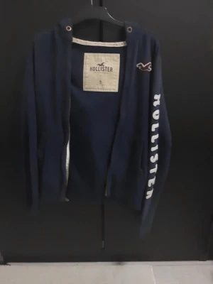 Mörkblå hoodie från Hollister - Mörkblå hoodie från Hollister med dragkedja, huva och vita tryckta bokstäver längs ena ärmen. Klassisk Hollister-logga på bröstet och snörning i huvan. Perfekt för en avslappnad och stilren look.