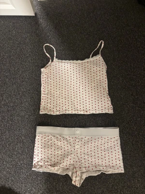 Vit pyjamas med röda hjärtan - Söt pyjamas med linne och shorts i vitt med små röda hjärtan över hela tyget. Linnet har tunna axelband och shortsen har bred resår i midjan samt dekorativa knappar framtill. Perfekt för mysiga kvällar hemma.