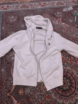 Vit hoodie från Polo Ralph Lauren - Snygg vit hoodie från Polo Ralph Lauren med dragkedja och huva. Klassisk design med två fickor framtill och den ikoniska blå loggan broderad på bröstet. Tillverkad i mjukt och bekvämt material, perfekt för en avslappnad stil.