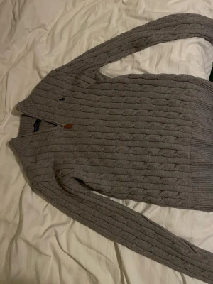 Grå kabelstickad half-sip Polo Ralph Lauren - Snygg grå kabelstickad tröja från Polo Ralph Lauren med dragkedja framtill och ribbade muddar. Klassisk design med liten broderad logga på bröstet. Perfekt till lager på lager och enkel att matcha med jeans. Ganska stora i storlek, kan passa L då det är lite stretch i tröjan