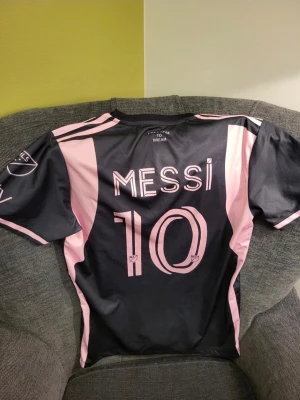 Inter Miami Messi matchtröja svart/rosa M - Snygg svart och rosa matchtröja från Inter Miami CF med Messi och nummer 10 på ryggen. Tröjan har klubbens officiella logga på bröstet och är tillverkad i ett lätt, andningsbart material som passar perfekt till fotbollsträning eller match.