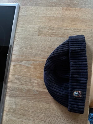 Mörkblå ribbstickad mössa Parajumpers - Säljer en mörkblå ribbstickad mössa från Parajumpers med uppvikt kant och klassisk PJS-logga framtill. Mössan är gjord i mjukt material och har en stilren, enkel design som passar perfekt till kalla dagar.