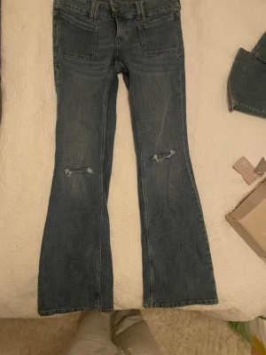 Mörkblå bootcut jeans med slitningar - Säljer ett par mörkblå bootcut jeans med slitna detaljer på knäna och dubbla knappar i midjan. Jeansen har stora framfickor och klassisk femficksdesign. Perfekta för en avslappnad och trendig look.