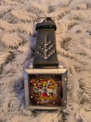 Ed Hardy klocka med tigerdesign - Säljer en fyrkantig klocka från Ed Hardy med en färgstark tiger på urtavlan och svart läderarmband med vit broderad detalj. Boetten är i silverfärgad metall och har markerade skruvar i hörnen för en cool look.