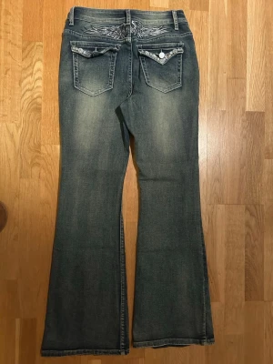 Blå bootcut jeans med broderi - Säljer ett par blå bootcut jeans med snyggt slitna detaljer och broderade fickor bak. Jeansen har vita kontrastsömmar och dekorativa knappar på bakfickorna. Perfekta för dig som gillar en avslappnad men ändå trendig stil.