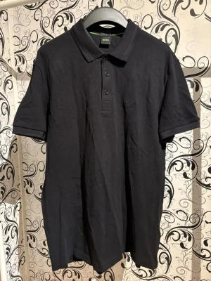 Svart Hugo Boss piketröja - Svart Hugo Boss piketröja i regular fit. Tillverkad i bomull med klassisk krage och knäppning framtill. Diskret broderad Hugo Boss-logotyp på bröstet. I mycket bra skick.