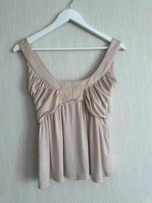 Beige omlott linnetopp med volang - Säljer en ljusbeige linnetopp med breda axelband och snygga volangdetaljer framtill. Toppen har omlottkänsla och ett mjukt, lätt glansigt material som ger en lyxig vibe. Perfekt för dig som vill ha något stilrent men ändå unikt.