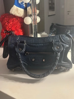 Mörkblå Balenciaga handväska - Säljer en mörkblå handväska från Balenciaga med klassisk city-modell. Väskan har silverfärgade nitar, dragkedja framtill och flätade handtag. Tillverkad i skinn med en cool, sliten look och detaljerade sömmar. Perfekt för dig som gillar statement-accessoarer.