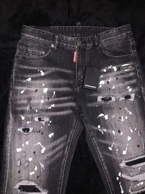 Svarta Dsquared2 jeans med färgstänk - Säljer ett par svarta jeans från Dsquared2 med vitmålade färgstänk och slitningar på benen. Jeansen har en smal passform, klassisk femficksdesign och en liten röd logotagg vid gylfen. Materialet är denim med stretch för extra komfort.