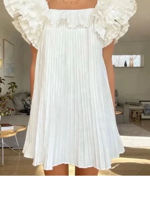 Vit plisserad kortklänning med volang - Supersöt vit kortklänning med plisserat tyg och stora volangdetaljer vid axlarna. Klänningen har en luftig passform och är ärmlös, perfekt för varma dagar. Ger en romantisk och trendig look.