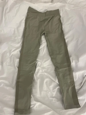 Olivgröna leggings med ribbade sidor - Snygga olivgröna leggings med bred midjeresår och ribbade detaljer längs sidorna. Tight passform och stretchigt material. Perfekta för dig som gillar stilrena och bekväma plagg till vardags eller träning.