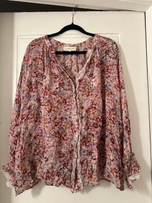 Blommig blus från By Malina - Ivey Blouse from By Malina - Säljer en luftig blommig blus från By Malina i rosa, lila och vita toner. Blusen har långa ärmar, volangdetaljer och spets längs knäppningen och ärmsluten. Materialet är tunt och flowy, perfekt för en romantisk look.