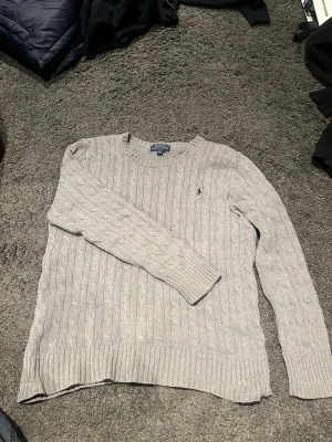 Ljusgrå kabelstickad tröja Polo Ralph Lauren - Kabelstickad ljusgrå tröja från Polo Ralph Lauren med rund halsringning och diskret broderad logga på bröstet. Tröjan har långa ärmar och ribbade muddar vid ärmslut och nederkant. Perfekt för lager på lager och klassisk stil.