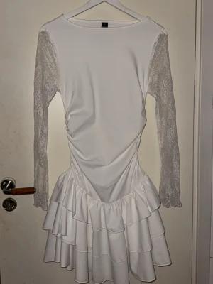 Vit volangklänning med spetsärmar - Säljer en vit kortklänning med långa spetsärmar och flera lager volanger i kjolen, fick hem klänningen idag men den passade tyvärr inte mig därför säljer den så helt ny. Klänningen har en figurnära passform 