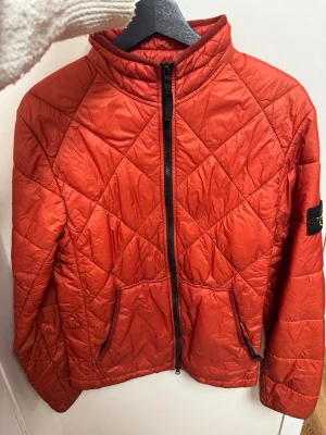 Röd pufferjacka från Stone Island - Säljer en röd pufferjacka från Stone Island med quiltat mönster och svart dragkedja framtill. Jackan har hög krage, två sidofickor med dragkedja och den klassiska Stone Island-loggan på ärmen. Materialet är glansigt och lätt, perfekt för kalla dagar. (Skulle säga att färgen  är orange/röd)